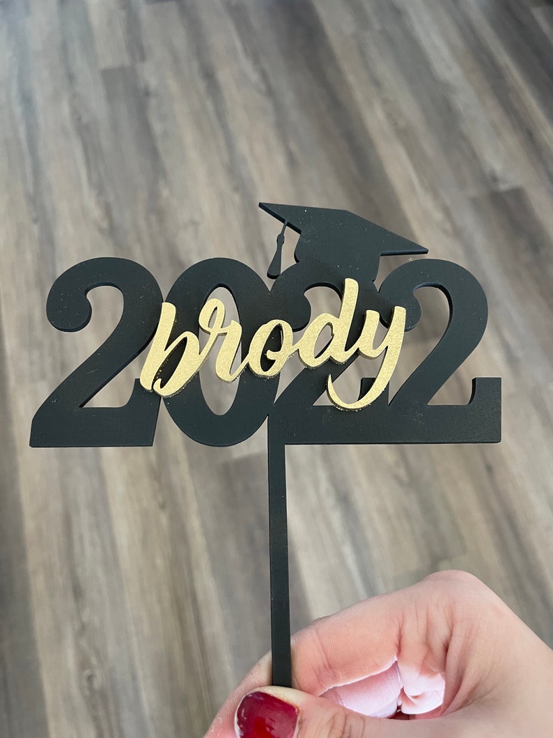 Pu&ograve; includere: Decorazione per torta di laurea nera e oro con l'anno 2022 e il nome "brody". La decorazione include una silhouette del cappello di laurea. La decorazione &egrave; tenuta da un sottile bastoncino nero.