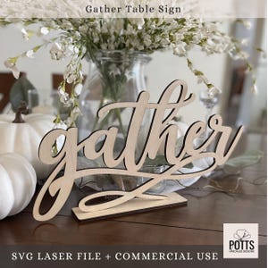 May include: A wooden "Gather" table sign in a cursive font, with a separate base for standing. The sign is light-colored wood with a dark outline. The text "Gather Table Sign" is at the top. The sign is on a table with white pumpkins and flowers.