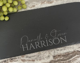 Puede incluir: Una tabla de servir rectangular de pizarra negra con bordes naturales y rugosos. La superficie está grabada con el texto Danielle & Steven HARRISON en una combinación de fuentes script y sans-serif. Un racimo de uvas verdes descansa en la esquina.