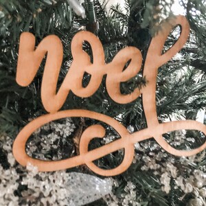May include: A wooden Christmas ornament with the word "Noel" written in cursive script. The ornament is hanging on a Christmas tree.
