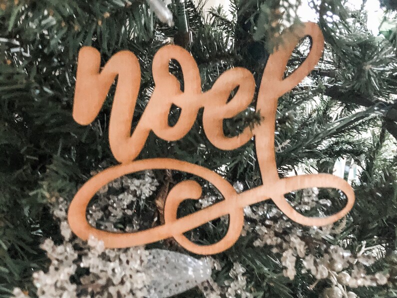 May include: A wooden Christmas ornament with the word "Noel" written in cursive script. The ornament is hanging on a Christmas tree.