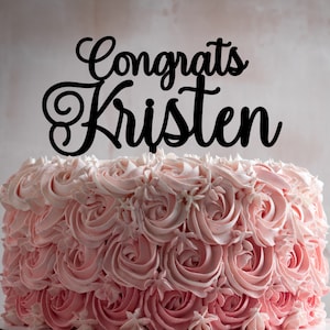 May include: A pink and white ombre cake decorated with buttercream frosting in a rose pattern. The cake is topped with a black acrylic cake topper that says "Congrats Kristen".