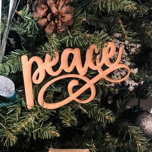 May include: A wooden sign with the word "peace" written in cursive script. The sign is hanging on a Christmas tree with pine needles and a brown pine cone.