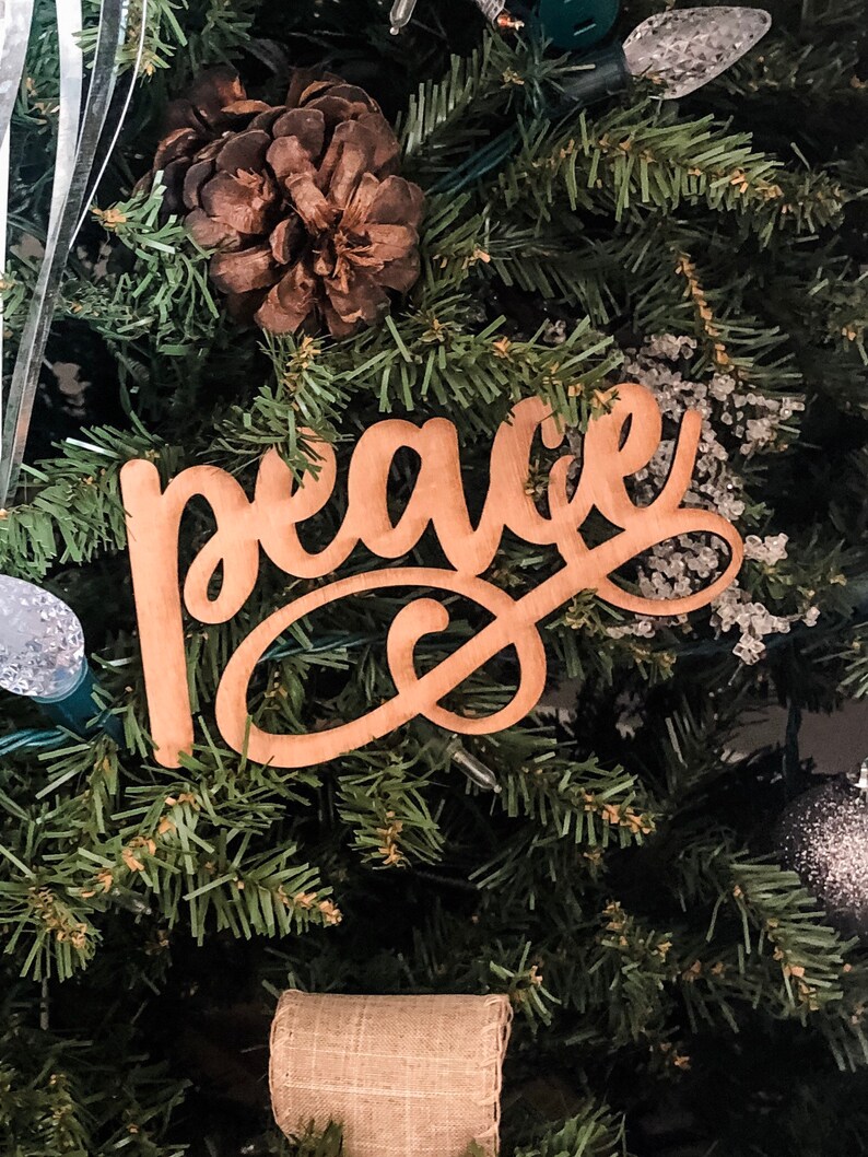 May include: A wooden sign with the word "peace" written in cursive script. The sign is hanging on a Christmas tree with pine needles and a brown pine cone.