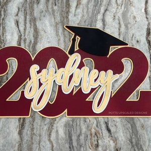 personalized graduation sign 2022 in maroon sydney in gold and a black graduation cap created for senior night gifts and graduation party decor by Potts Upscaled Designs LLC