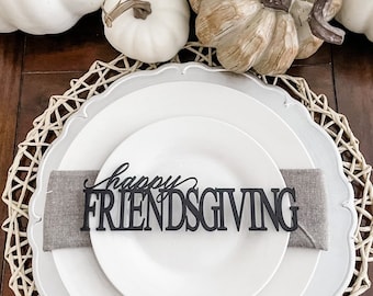 May include: A white dinner plate with a gray napkin and a black wooden sign that says "Happy Friendsgiving" on a woven placemat.