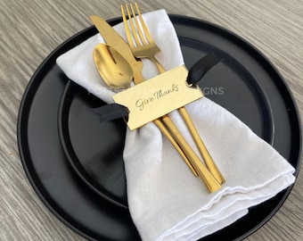 May include: A gold-toned stainless steel fork, knife, and spoon set resting on a white cloth napkin. A rectangular gold acrylic tag with the engraved text Give Thanks is secured to the cutlery with a thin black ribbon, all placed on a black plate.