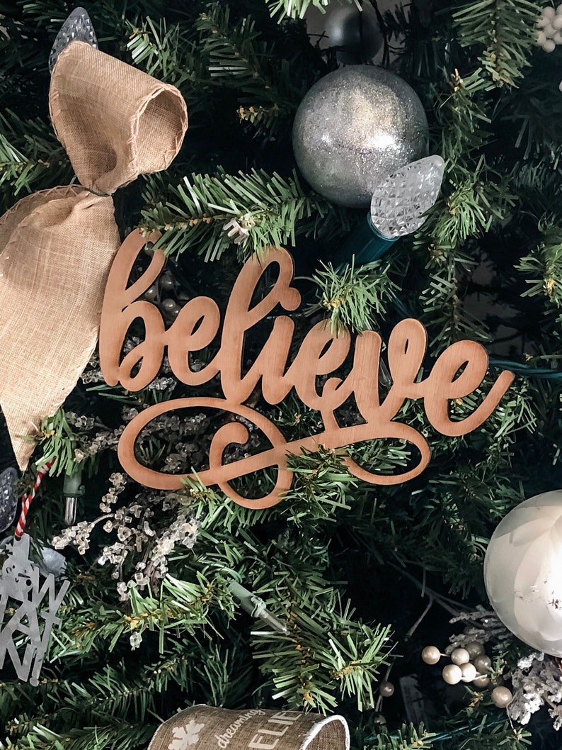 May include: A wooden sign with the word "believe" in cursive script, hanging on a Christmas tree decorated with burlap ribbon, silver ornaments, and white berry sprigs.