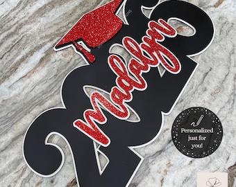 Puede incluir: Un letrero de graduación negro con el año 2026, un birrete rojo con purpurina y el nombre "Madalyn" en purpurina roja. El letrero incluye el texto "Graduation Sign - Deluxe - 2026" y "¡Personalizado solo para ti!"