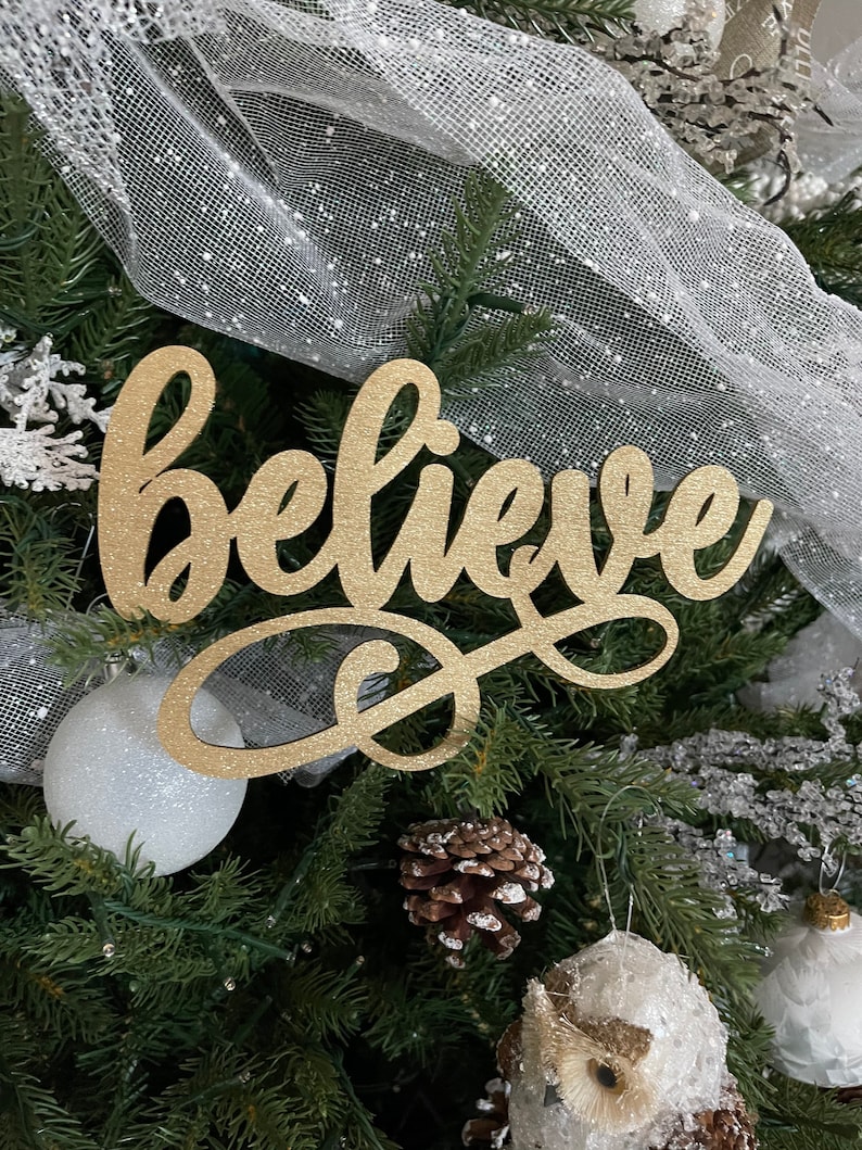 May include: A gold glitter "believe" sign ornament hanging on a Christmas tree with white and silver decorations.