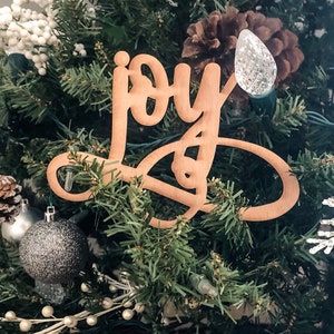 May include: A wooden Christmas tree ornament with the word "joy" written in cursive script. The ornament is hanging on a green Christmas tree with white lights and other decorations.