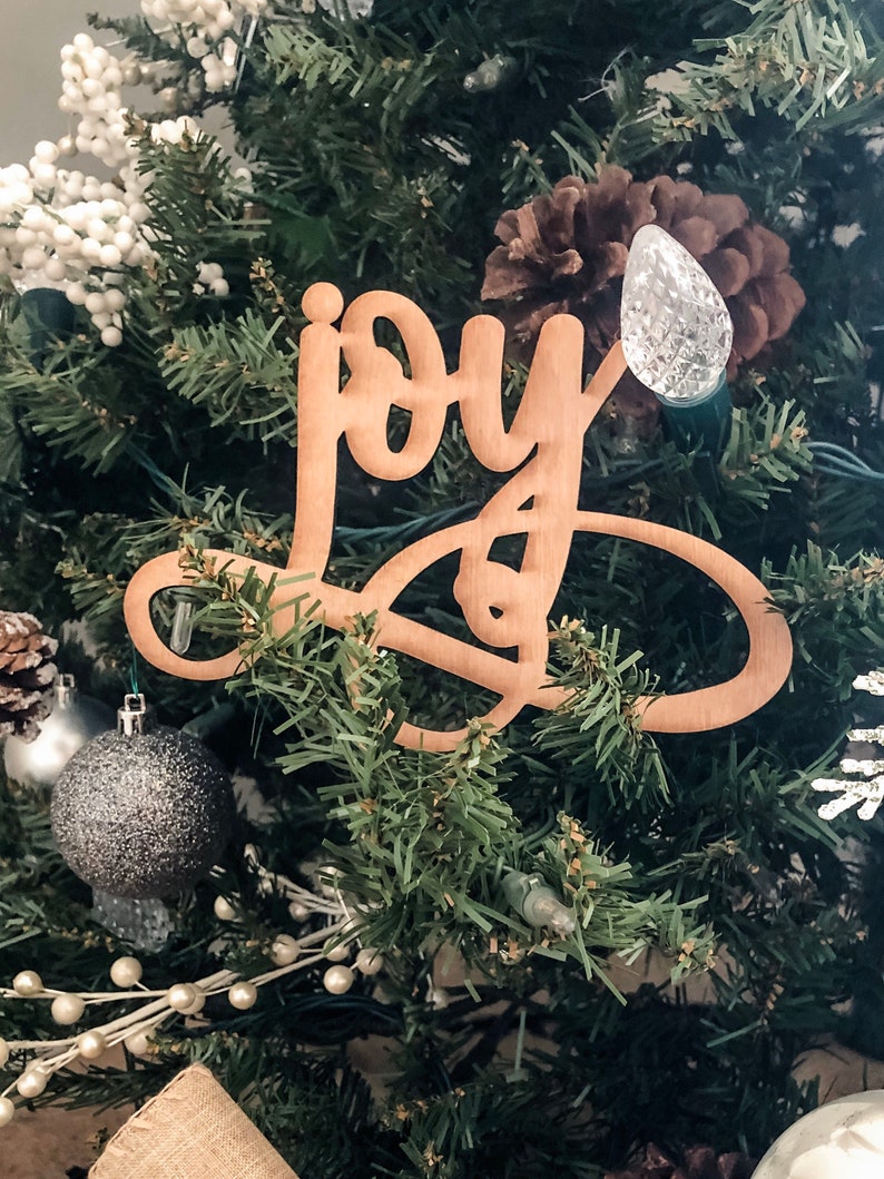 May include: A wooden Christmas tree ornament with the word "joy" written in cursive script. The ornament is hanging on a green Christmas tree with white lights and other decorations.