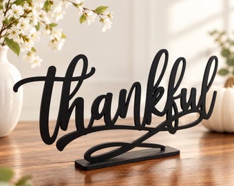 May include: A black tabletop sign featuring the word thankful written in a cursive script font. The sign is constructed from a flat material and stands upright on a rectangular base, displaying a smooth matte finish across the entire surface.