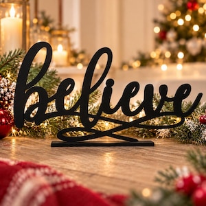 May include: A black, cursive-style tabletop sign featuring the word believe in a flowing script font. The sign is constructed from a flat, solid material and stands on a rectangular base, showcasing a clean, dark silhouette against a festive setting.