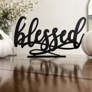 May include: Black metal sign with the word "blessed" in a cursive font. The sign is sitting on a wooden table with white pumpkins and a vase of white flowers in the background.