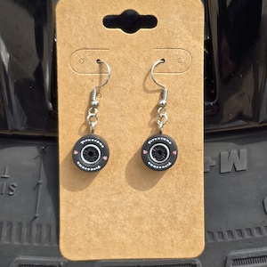 May include: A pair of black and silver earrings shaped like miniature tires. The earrings have a silver hook and a small pink heart on each tire. The tires have the word "Firestone" printed on them.