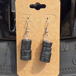 May include: A pair of black tire stack earrings with silver hooks. The earrings are displayed on a brown card.