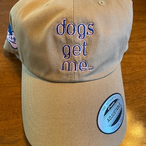 May include: A tan baseball cap with an embroidered design that says "dogs get me." The cap has an adjustable strap with a silver tag that says "The Authentic Yupoong Adjustable Since 1974."