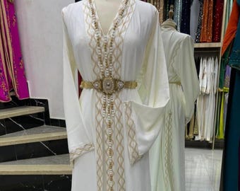 May include: A white, long-sleeved dress with gold embroidery and a matching belt. The dress features intricate detailing along the front, sleeves, and hem. The dress is displayed on a mannequin, with a mirror reflecting the back of the dress.