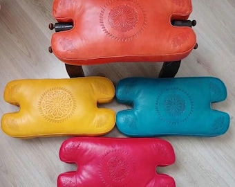 May include: A collection of colorful leather ottomans in orange, yellow, teal, and red. Each ottoman has a unique shape with a central embossed floral design. One ottoman is on a wooden stool.