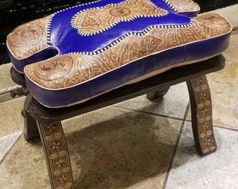 May include: A handcrafted, decorative stool with a blue and tan leather seat. The seat features intricate embossed patterns and is supported by a dark wooden frame. The legs are also decorated with carved designs, creating a unique and artistic piece.