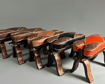 May include: A row of handcrafted wooden stools with leather seats. The seats are in various colors, including brown, black, and orange, and feature intricate patterned designs. The stools have curved legs and decorative metal accents.
