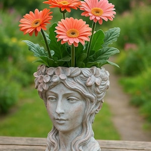 Puede incluir: Una maceta decorativa con forma de cabeza de mujer, llena de vibrantes margaritas gerberas naranjas y rosas. La maceta es de color gris claro con detalles florales alrededor de la cabeza. El fondo es un jardín verde borroso.