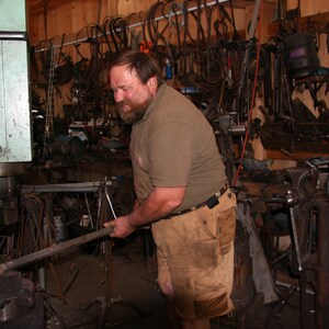 May include: A man in a brown shirt and tan pants uses a large metal press to shape a metal rod. The workshop is filled with tools and equipment.