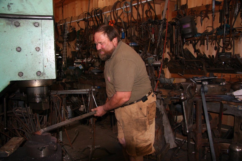 May include: A man in a brown shirt and tan pants uses a large metal press to shape a metal rod. The workshop is filled with tools and equipment.