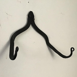 May include: A black, wrought iron wall hook with a distinctive, curved design. The hook features two downward-facing hooks and two upward-curving arms, creating a decorative and functional piece for hanging items.