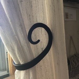 May include: Black metal curtain tieback with a decorative swirl design. The tieback is holding a white sheer curtain.