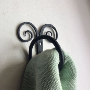 May include: A black wrought iron towel ring with decorative scrollwork and a circular ring. A light green knitted towel is hanging from the ring. The towel ring is mounted on a white wall.