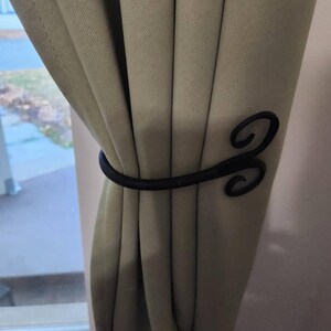 May include: Black metal curtain tieback with a decorative swirl design. The tieback is holding back a pair of beige curtains.