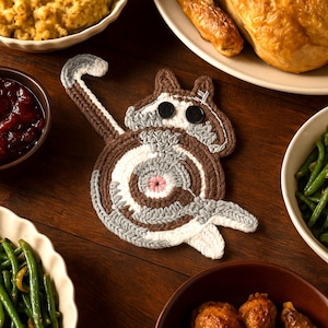 Puede incluir: Un posavasos de ganchillo hecho a mano con forma de ardilla en marrón, blanco y gris. El posavasos presenta un diseño en espiral para el cuerpo y una cola. Se utilizan dos botones negros para los ojos. El posavasos está colocado sobre una mesa de madera con comida.