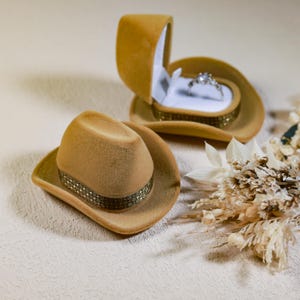 Könnte beinhalten: Zwei Miniatur-Ringboxen in Form von Cowboyhüten in Beige, eine davon geöffnet, um einen Diamantring zu zeigen. Die Hüte haben ein dekoratives Band um den Sockel. Ein kleiner Strauß Trockenblumen befindet sich im Hintergrund. Die Ringbox ist einzigartig.