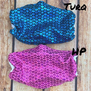 May include: Two face masks with a mermaid scale pattern. The top mask is turquoise and the bottom mask is pink.