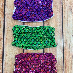 May include: Three face masks with a mermaid scale pattern. The masks are purple, green, and pink with glitter accents.