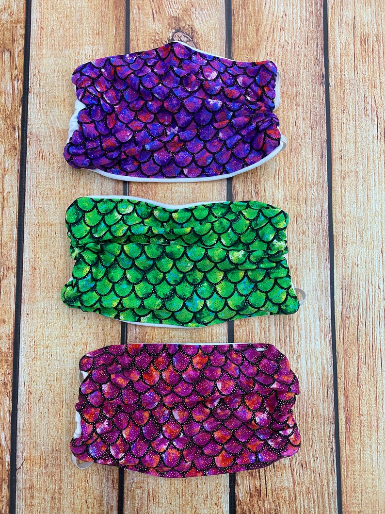 May include: Three face masks with a mermaid scale pattern. The masks are purple, green, and pink with glitter accents.