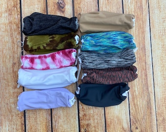 May include: A stack of 10 colorful fabric face masks with different patterns, including tie-dye, camouflage, and stripes. The masks are arranged in two columns on a wooden background.