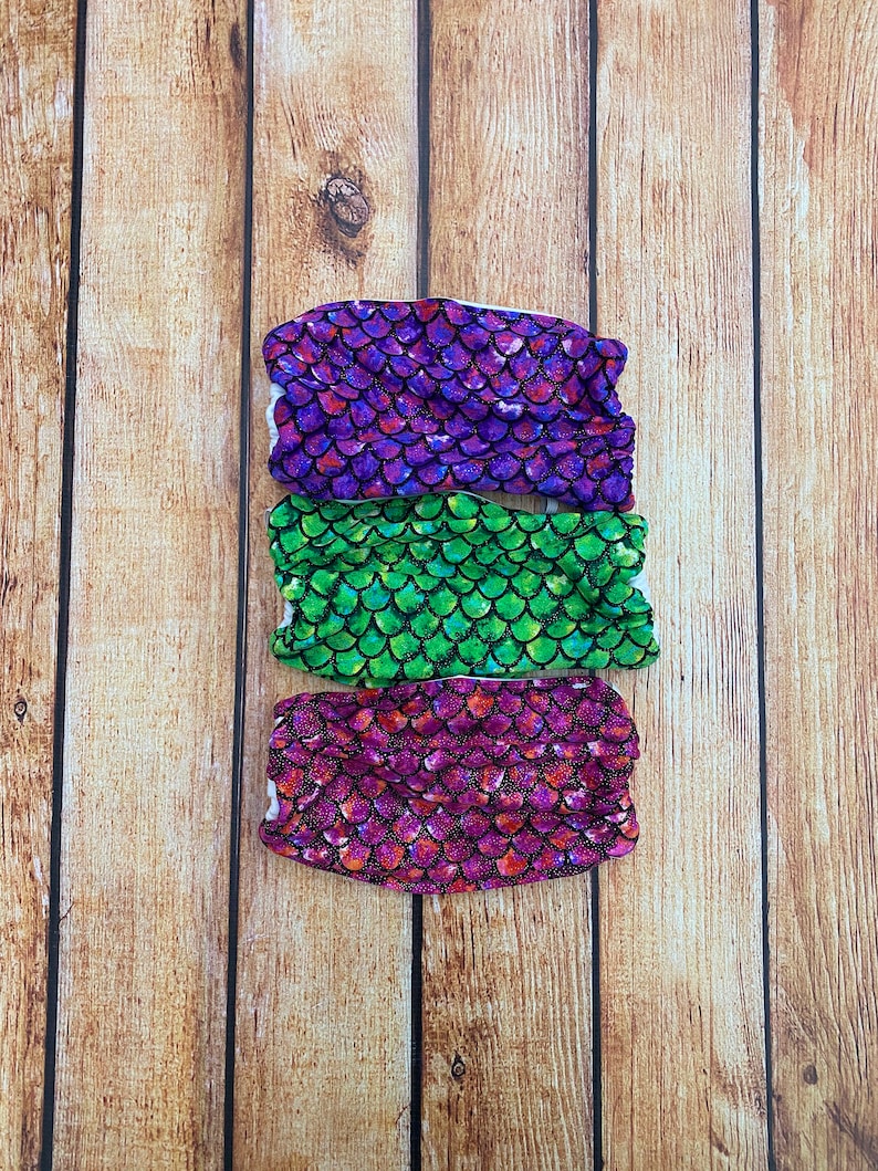 May include: Three fabric face masks with a mermaid scale pattern. The masks are purple, green, and pink.