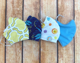 May include: Four fabric face masks with different patterns. The masks are light blue, white with colorful circles, navy blue with a floral pattern, and yellow with a geometric pattern.