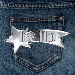 May include: A silver star-shaped pin with the text "by flo" on a blue denim background.
