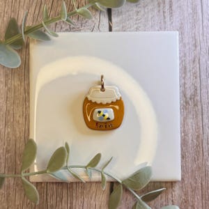 May include: A gold-toned metal pendant shaped like a honey jar with a white lid, a central label featuring a small bee illustration, and the name "leia" written in lowercase script. The pendant includes a small metal loop at the top for hanging.