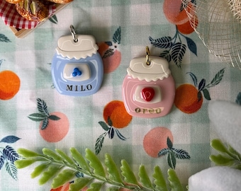 May include: Two jar-shaped charms with white lids and metal loops. The light blue charm features a blue berry accent and the text MILO. The pink charm features a red strawberry accent and the text OREO. Both charms have a smooth, glossy finish.