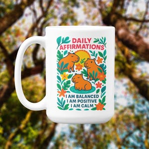 May include: White ceramic mug featuring a colorful illustration of capybaras with the text "DAILY AFFIRMATIONS." The design includes the affirmations "I AM BALANCED, I AM POSITIVE, I AM CALM" with floral accents. The mug has a large handle.
