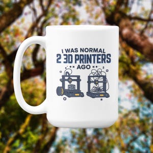 Könnte beinhalten: Weißer Keramikbecher mit großem Henkel. Der Becher zeigt den Text "I WAS NORMAL 2 3D PRINTERS AGO" in Blau, zusammen mit Illustrationen von 3D-Druckern. Der Hintergrund ist verschwommen mit Grün- und Brauntönen.