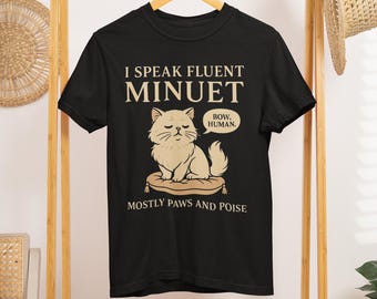 Może przedstawiać: Czarny t-shirt z napisem "I Speak Fluent Minuet" nad rysunkowym kotem na poduszce. Kot ma dymek z napisem "Bow, Human". Pod kotem znajduje się napis "Mostly Paws and Poise".