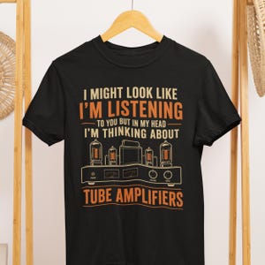 May include: A black short-sleeved t-shirt featuring orange and cream text that reads I MIGHT LOOK LIKE I'M LISTENING TO YOU BUT IN MY HEAD I'M THINKING ABOUT TUBE AMPLIFIERS above a graphic illustration of a vintage vacuum tube audio amplifier.