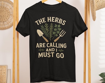 May include: Black t-shirt with the text "THE HERBS ARE CALLING AND I MUST GO" in white and green. The design includes illustrations of gardening tools and herbs, perfect for gardening enthusiasts.