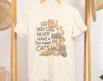 May include: Cream-colored t-shirt featuring a cartoon illustration of several cats in various poses. The text "YOU CAN NEVER HAVE TOO MANY CATS" is printed in brown. The shirt is hanging on a wooden clothing rack.